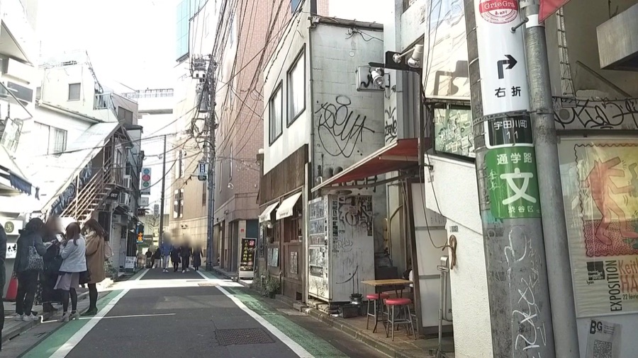 A narrow school route in Shibuya, captured in 2018, with green edge lines, a school-route sign, and pedestrians